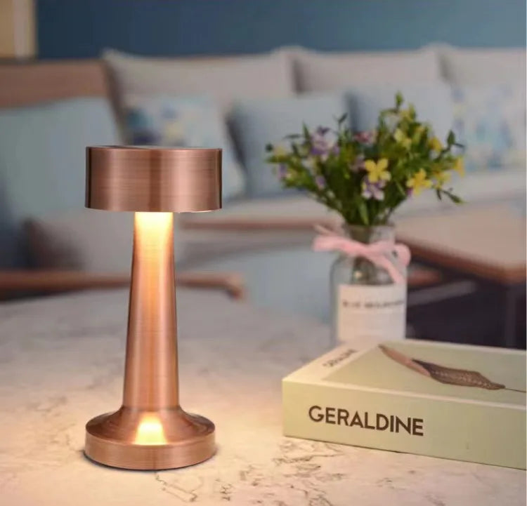 Lampe de table cuivrée allumée à côté d’un livre et d’un vase.